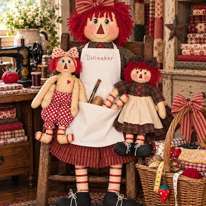May include: A large rag doll with red yarn hair and a striped bow, wearing a white apron embroidered with the word Dollmaker, sits on a wooden chair. Two smaller rag dolls with red hair and matching outfits are positioned on either side of the large doll.