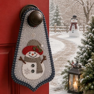 May include: A gray felt door hanger featuring a snowman applique with a red hat, brown scarf, and two black buttons. The hanger has a dark blue stitched border and is suspended from a round bronze-colored door knob on a red door.