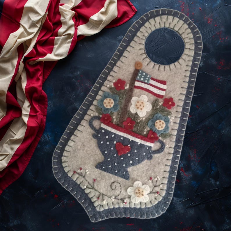 May include: A felt door hanger featuring a patriotic design. It showcases a blue polka-dotted vase with red and white flowers, an American flag, and a heart. The border is a light gray with a dark blue stitched edge.
