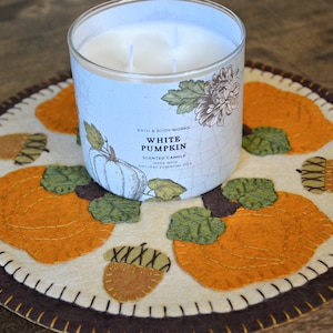 May include: A white candle with a label that reads "Bath & Body Works White Pumpkin Scented Candle Made with Natural Essential Oils" is sitting on a cream-colored felt coaster with an orange, brown, and green pumpkin design.