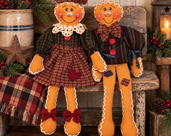May include: Two plush gingerbread dolls with painted faces and stitched white icing details. The left doll wears a plaid dress with a lace collar and hair bow. The right doll wears a plaid shirt with a bow tie. Both have tan fabric bodies.