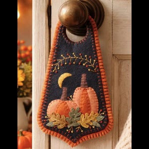 May include: A felt door hanger with a navy blue background and an orange border. The design features two orange pumpkins, a crescent moon, and fall foliage. The hanger is attached to a brass doorknob.