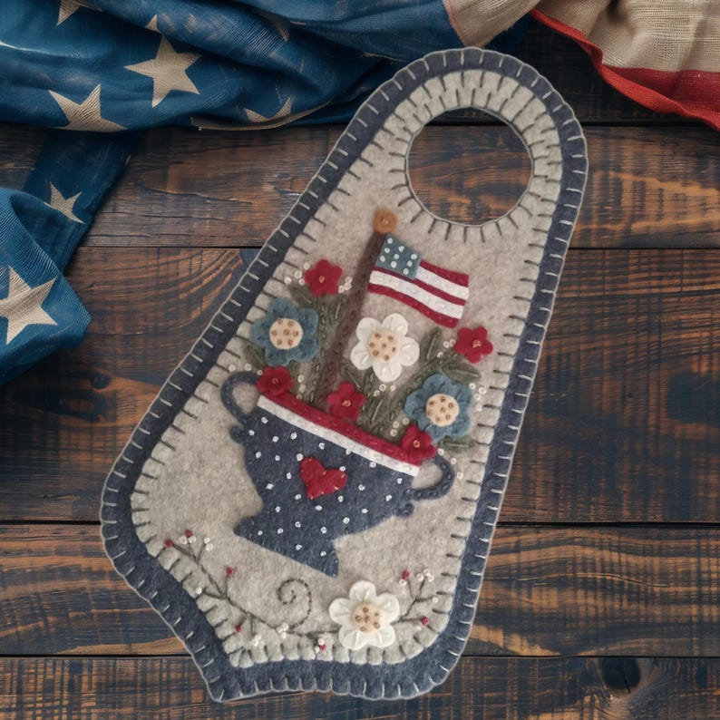 May include: A felt door hanger with a patriotic theme. It features a blue vase with white polka dots, red and white flowers, and an American flag. The background is a light gray with a dark blue border and a hole at the top for hanging.