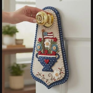 May include: A patriotic door hanger featuring a felt design with a vase of flowers, an American flag, and a blue border. The vase has a red heart and blue polka dots. The door hanger is attached to a gold doorknob.