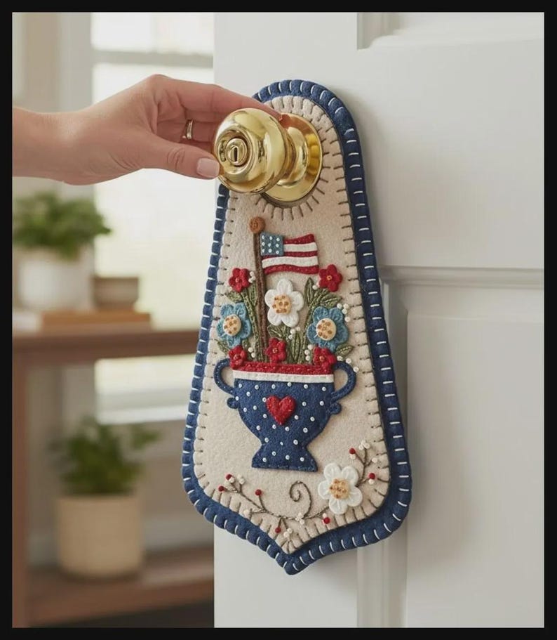 May include: A patriotic door hanger featuring a felt design with a vase of flowers, an American flag, and a blue border. The vase has a red heart and blue polka dots. The door hanger is attached to a gold doorknob.