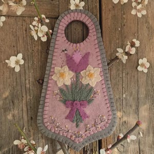 May include: A handmade felt door hanger in shades of pink and gray, featuring a floral bouquet with a purple tulip, white daffodils, and a purple bow. A small bee is stitched above the flowers. The hanger is decorated with small white and yellow beads.