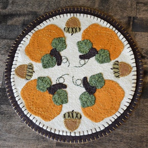 Peut inclure: Un chemin de table rond en feutre blanc avec une bordure en feutre brun. Le chemin de table présente quatre citrouilles orange avec des feuilles vertes et des tiges brunes, et quatre glands bruns avec un X blanc sur chacun.