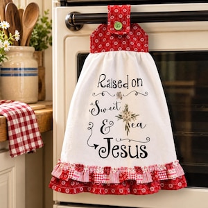 May include: A white hanging kitchen towel with a red floral patterned top and a matching ruffled hem. The towel features the black text "Raised on Sweet Tea & Jesus" with a small floral cross graphic, secured by a green button on the oven handle.