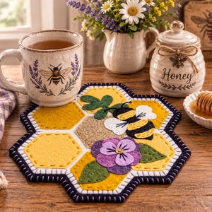 May include: A hexagonal felt coaster featuring a yellow honeycomb pattern, a black and yellow bee appliqué, a purple pansy flower, and green leaves. The coaster has a dark navy blue stitched border and is displayed with a bee-themed mug and honey pot.