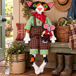 May include: A handcrafted fabric doll featuring a calico cat head with green eyes and long whiskers, wearing a green floral shirt, brown plaid overalls, and a red bandana. The cat holds a small grey mouse doll with pink ears and a red bandana.