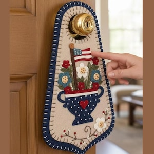 May include: A patriotic door hanger featuring a felt design with a vase of flowers and an American flag. The door hanger has a cream-colored background with a blue border and decorative stitching. A hand is pointing at the flag.