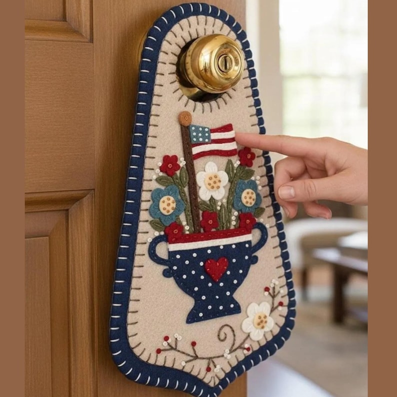 May include: A patriotic door hanger featuring a felt design with a vase of flowers and an American flag. The door hanger has a cream-colored background with a blue border and decorative stitching. A hand is pointing at the flag.