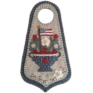 May include: A felt door hanger in a patriotic theme. The gray felt has a blue stitched border and a large hole at the top. A blue and white polka-dotted vase with a red heart holds flowers and an American flag.