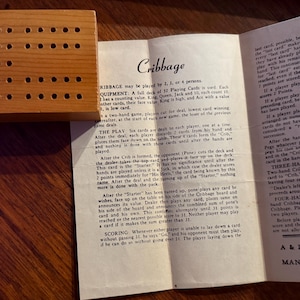 Könnte beinhalten: Ein hölzernes Cribbage-Spielbrett mit mehreren Löchern liegt neben einem offenen Anleitungsblatt. Das Blatt beschreibt die Regeln von Cribbage, einschließlich der Punktevergabe und des Spielverlaufs. Das Spiel ist für 2, 3 oder 4 Spieler.