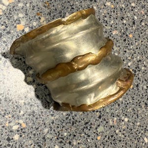 May include: A silver-colored, sculpted metal cuff bracelet with a textured surface. The bracelet is accented with a gold-colored trim around the edges and a band in the middle. The cuff has a unique, artistic design.