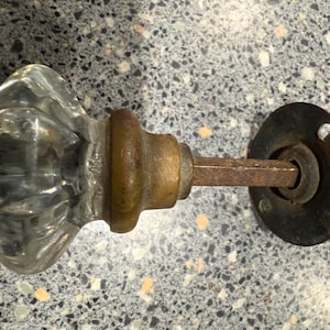 Puede incluir: Pomo de puerta de cristal antiguo con un mango de cristal transparente y acanalado y herrajes de latón. El pomo presenta un diseño clásico con una placa trasera redonda y un husillo de metal oxidado. Un artículo de decoración vintage.