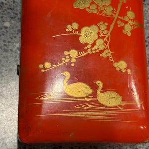 Puede incluir: Caja rectangular roja con una rama floral dorada y dos patos dorados nadando sobre una superficie de agua dorada. La caja tiene un aspecto ligeramente desgastado.