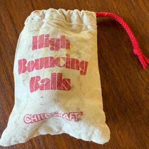 May include: A beige canvas drawstring bag with the words "High Bouncing Balls" printed in red. The bag also has the word "Childcraft" printed in red. A red drawstring is attached to the top of the bag.