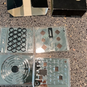 Peut inclure: Un ensemble de quatre jeux de patience argentés dans des étuis en plastique transparent. Les jeux sont étiquetés "Geduldspiele Set" et comprennent un labyrinthe, un jeu de pions et deux autres jeux avec des formes géométriques.