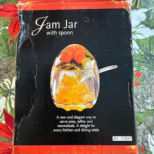 Può includere: Una scatola nera e rossa con la scritta "Jam Jar with spoon". La scatola presenta un'illustrazione di un barattolo di marmellata di vetro con un cucchiaio, pieno di marmellata arancione e sormontato da frutta arancione. Il testo recita: "Un modo nuovo ed elegante per servire marmellate, gelatine e marmellate."