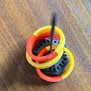 May include: Three colorful bracelets in yellow, orange, and red rest on a dark gray, geometric-patterned object. A dark, thin object is inserted through the bracelets. The bracelets are on a wooden surface.