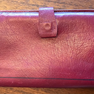 May include: A burgundy leather wallet with a rectangular shape. The wallet has a flap closure with a button and a black border. The leather has a textured appearance.