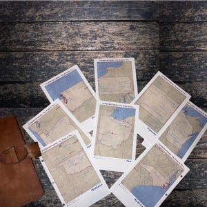 Puede incluir: Una colección de impresiones de mapas con varios estados de EE. UU., dispuestas en un patrón circular. Cada impresión tiene una estética vintage con un fondo de cuadrícula y el texto "SUPERNATURAL: The Road So Far..." en la parte superior. Un diario de cuero marrón es visible a la izquierda.