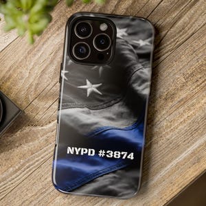 May include: A smartphone case featuring a black and white American flag design with a blue stripe. The case has the text "NYPD #3874" in white. The case is on a wooden surface.