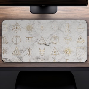 May include: Rectangular desk mat with a white marble pattern and gold geometric symbols. The symbols include triangles, sunbursts, and other abstract shapes. The mat has a black border and is designed to protect a desk surface.