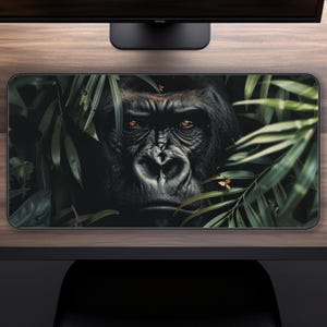 May include: Rectangular desk mat featuring a close-up of a gorilla's face surrounded by green foliage. The gorilla has dark fur and intense eyes. The mat is designed to protect the desk surface.
