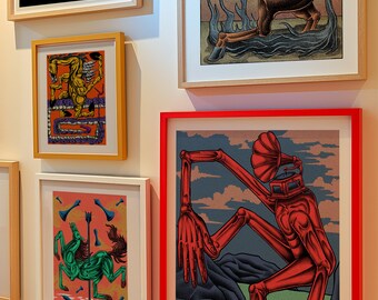 May include: A collection of framed art prints featuring various illustrations. One print depicts a red humanoid figure with a large ear-like structure on its head, another shows a green horse-like creature, and another features a brown horse with flames. The prints are displayed on a white wall.