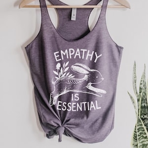 May include: A heather purple tank top with a white graphic of a rabbit and the words "EMPATHY IS ESSENTIAL". The tank top is hanging on a wooden hanger against a white wall. A green plant is visible on the right.