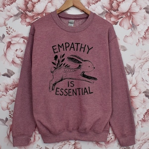 May include: A maroon sweatshirt with the words "EMPATHY IS ESSENTIAL" and a black illustration of a rabbit. The sweatshirt is hanging on a wooden hanger against a floral background.