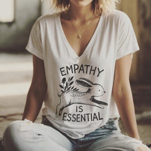 May include: A woman wearing a white V-neck t-shirt with the text "EMPATHY IS ESSENTIAL" and a graphic of a rabbit with leaves. She is sitting on the floor in a room with large windows. She is wearing a gold necklace and blue jeans.