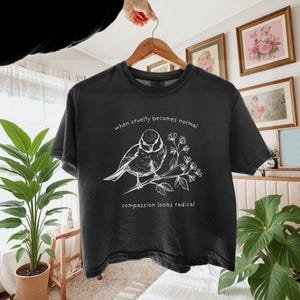 May include: A black short-sleeve t-shirt featuring a white line art illustration of a bird perched on a flowering branch. The text "when cruelty becomes normal" is printed above the bird, and "compassion looks radical" is printed below.