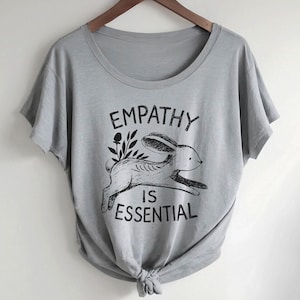 May include: A gray t-shirt with a round neck and short sleeves. The shirt features a black graphic of a running rabbit with the words "EMPATHY IS ESSENTIAL" printed above and below the rabbit. The shirt is knotted at the bottom.