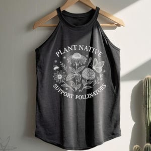 May include: Dark gray halter top with the text "PLANT NATIVE SUPPORT POLLINATORS" in a circular design with floral and butterfly illustrations. The tank top is hanging on a wooden hanger against a neutral wall, with a cactus in the background.