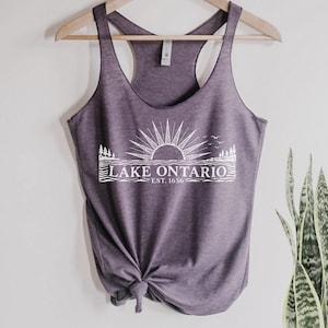 May include: A heathered purple racerback tank top featuring a white graphic print of a sun rising over water with pine trees and birds. The text reads LAKE ONTARIO EST. 1656. The garment is displayed on a wooden hanger with a knot tied at the bottom.