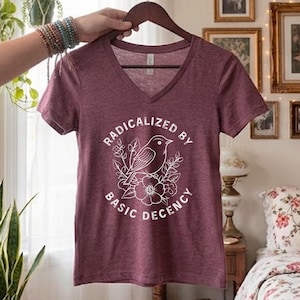 May include: A heathered maroon V-neck t-shirt featuring a white graphic print of a bird perched on a flower branch. The text "RADICALIZED BY BASIC DECENCY" is printed in a circular arc around the central bird and floral illustration.