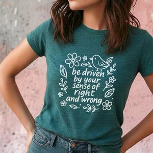 May include: Teal crew neck t-shirt with white floral and bird graphic. The text reads "be driven by your sense of right and wrong." The shirt is made of a soft material and is paired with blue jeans.