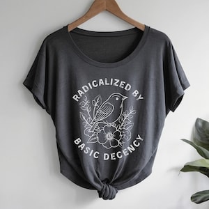 May include: A dark gray short-sleeve t-shirt with a wide scoop neckline and a knotted hem. The front features a white graphic of a bird perched on flowers with the text RADICALIZED BY BASIC DECENCY printed in a circular arrangement around it.