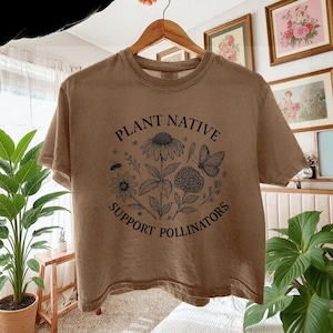 May include: A brown short-sleeve t-shirt featuring a black line-art graphic of various flowers, a butterfly, and a bee. The text "PLANT NATIVE" is printed in an arch above the illustration, and "SUPPORT POLLINATORS" is printed in an arch below.