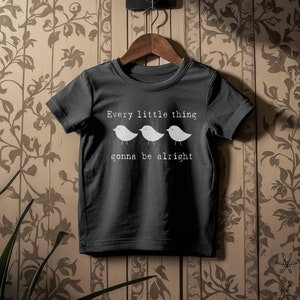 May include: Dark gray t-shirt with the text "Every little thing gonna be alright" and three white bird illustrations. The shirt is hanging on a wooden hanger against a patterned background.