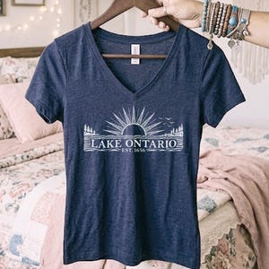 May include: A navy blue V-neck t-shirt featuring a white graphic print of a sun rising over Lake Ontario with pine trees and birds. The text reads LAKE ONTARIO EST. 1656. The shirt is displayed on a wooden hanger against a soft, neutral background.