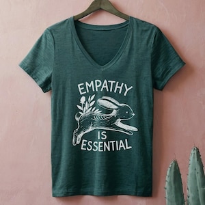 May include: A dark green V-neck t-shirt with the words "EMPATHY IS ESSENTIAL" in white, along with a white graphic of a running rabbit. The shirt is hanging on a wooden hanger against a pink background.