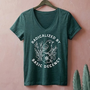 May include: A dark green V-neck t-shirt with the text "RADICALIZED BY BASIC DECENCY" surrounding a white illustration of a bird perched on flowers. The shirt is hanging on a wooden hanger.