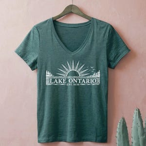 May include: A dark green V-neck t-shirt featuring a white graphic print of a sun rising over water with pine trees on the sides. The text reads LAKE ONTARIO EST. 1656. The shirt is displayed on a hanger against a solid light pink background.