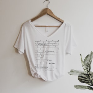 May include: A white V-neck t-shirt with a black brick wall design. The design includes the phrases "IT FEELS LIKE LOVE", "GOT THE RADIO ON", "AND IT'S", "ALL THAT WE NEED", "THERE'S A WHISTLETON BEAT", and "AND IT FEELS LIKE LOVE". The shirt hangs on a wooden hanger.