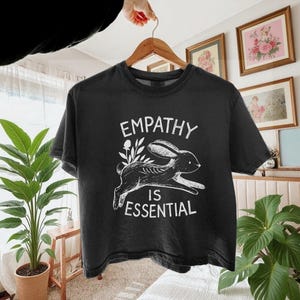 May include: Black t-shirt with the words "EMPATHY IS ESSENTIAL" in white. The design includes a running rabbit skeleton with floral accents. The shirt is hanging on a wooden hanger.