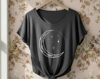 Puede incluir: Una camiseta gris oscuro de manga corta con un gráfico de líneas blancas de una luna creciente decorada con patrones de hojas botánicas y rodeada de pequeñas estrellas y puntos, con un detalle de nudo en el dobladillo central.
